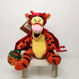 May include: A plush Tigger toy dressed in a devil costume, complete with a red cape and horns. He holds a small pumpkin-shaped treat bag and a red trident. The toy is orange with black stripes and a yellow belly.