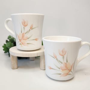 May include: Two white ceramic mugs with peach-colored floral designs. The mugs have curved handles and are displayed on a small, round, wooden stand. The floral design includes peach-colored flowers and green leaves.