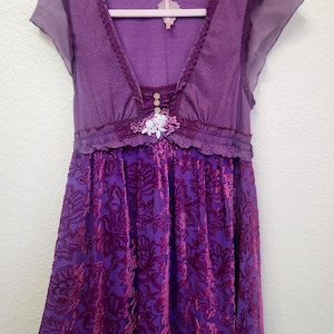 May include: A purple dress with a V-neckline and short sleeves. The bodice is a solid purple, while the skirt features a floral velvet pattern. The dress is embellished with lace and a decorative floral accent.