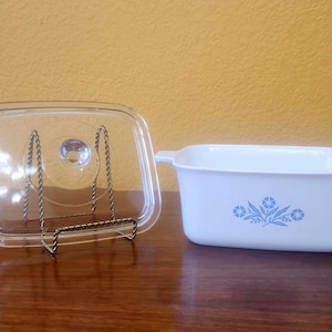 May include: A white rectangular glass baking dish with two side handles and a blue floral pattern on the front. Beside it stands a clear glass lid, supported by a metal display stand, featuring a central circular knob handle.