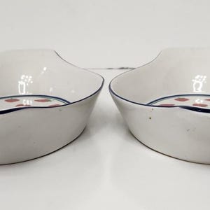 May include: Two white ceramic serving dishes with a navy blue trim and a pattern of red dots. The dishes have a unique shape with a curved rim and a slightly raised handle area.