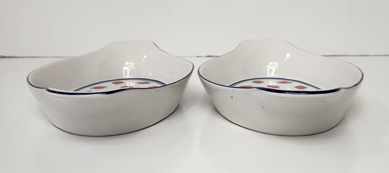 May include: Two white ceramic serving dishes with a navy blue trim and a pattern of red dots. The dishes have a unique shape with a curved rim and a slightly raised handle area.