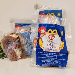 May include: A Ty Beanie Baby plush toy in a clear plastic bag with a McDonald's Happy Meal packaging. The toy is a brown squirrel with a white belly. The packaging is blue and yellow with the Ty Beanie Baby logo and the words "The Squiggle" and "Nuts".