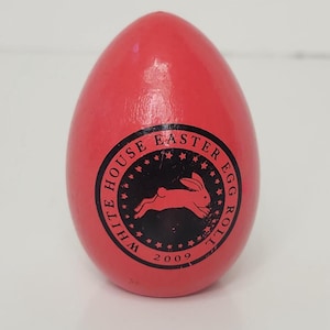 Puede incluir: Un huevo de madera rojo con un emblema circular negro con un conejo corriendo y el texto "WHITE HOUSE EASTER EGG ROLL 2009". Este huevo decorativo es probablemente para la Pascua.