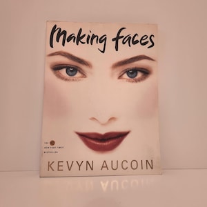 May include: A book titled "Making Faces" by Kevyn Aucoin. The cover features a close-up of a face with blue eyes and dark red lipstick. The book is a New York Times bestseller.