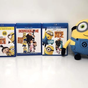 May include: Three Blu-ray movie cases for "Despicable Me" and a plush Minion toy. The movie cases are blue with colorful graphics of the Minions and characters. The plush toy is yellow and blue, resembling a Minion character.