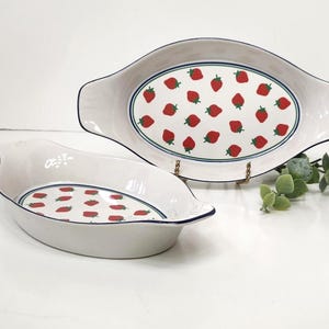 May include: Two white ceramic oval baking dishes. The dishes have a blue trim and a pattern of red strawberries with green stems. The larger dish is on a gold stand. The smaller dish is in front of the larger dish.