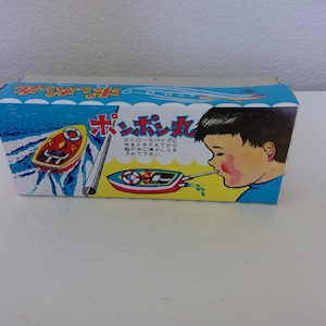 May include: A vintage toy box featuring a cartoon illustration of a boy blowing into a toy boat. The box is primarily blue and white, with Japanese text and images of a red and yellow boat. The box is rectangular.