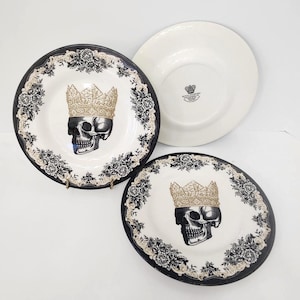 May include: White ceramic plates with a black and cream floral border. Two plates feature a skull wearing a gold crown. One plain white plate is also visible. The plates have a gothic or edgy aesthetic.