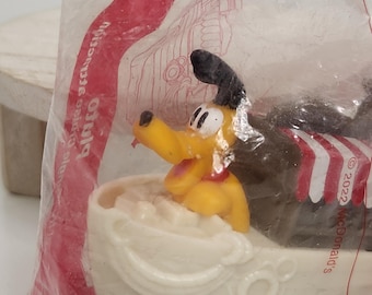 May include: A yellow Pluto figurine in a white boat with red and white striped accents. The toy is in a clear plastic bag. The boat is on a blue base. The text "© 2022 McDonald's" is visible.