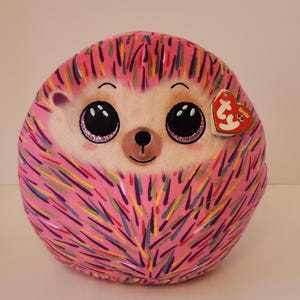 May include: A round, pink plush hedgehog toy with a smiling face, large glittery eyes, and a small nose. The hedgehog's body is covered in colorful, stitched lines. A red heart-shaped tag is attached to the side.