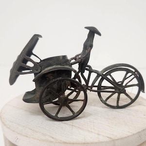 May include: A miniature antique bicycle figurine made of dark metal. The bicycle has two large wheels, a seat, and a driver figure. The bicycle is sitting on a light-colored, round wooden surface.