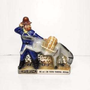 May include: A ceramic Ezra Brooks figurine depicting a man in a blue coat and brown hat with a donkey. The figurine has the text "ME and JIM FOUND TONOPAH, NEVADA" on the base. The man has a beard and is touching his hat.