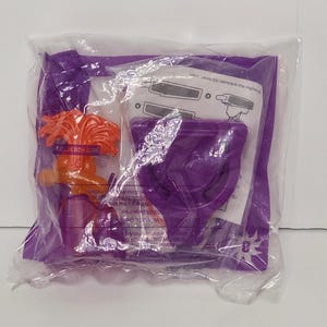May include: A sealed clear plastic bag containing a purple toy and an orange toy figure. The purple toy is a cup-like shape. The orange figure has a round head and arms. The bag has printed text and a number 3.