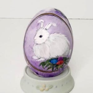 May include: A decorative Easter egg with a hand-painted white rabbit on a lavender background. The egg is set on a white ceramic stand with floral accents, creating a charming springtime display.