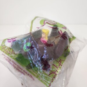 May include: A sealed plastic bag containing a small toy figurine. The figurine is of a blonde woman wearing a pink dress. The bag has green and purple accents and text that reads "Tulbag is not a toy. Please discard, Made in China. © 1998 Burger King Corporation."