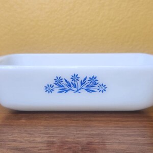 May include: A rectangular, white casserole dish with a blue floral design. The dish has a slightly flared shape and small handles on each end. The floral pattern is centered on the front of the dish.