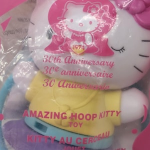 May include: A Hello Kitty 30th Anniversary toy in a plastic package. The toy is white with a pink bow and a yellow top. The text "AMAZING HOOP KITTY TOY" is printed on the toy. The text "Hello Kitty" and "1974" are also visible.