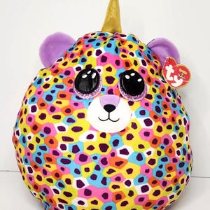 May include: A colorful plush toy in the shape of a unicorn with a spotted pattern. The toy features a gold horn, purple ears, and large black eyes with glitter accents. The plush has a white face with a small black nose and mouth.