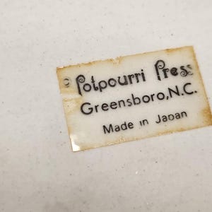 May include: Close-up of a white surface with a label that reads "Potpourri Press Greensboro, N.C. Made in Japan". The label is rectangular with a slightly aged appearance, and the text is in black.