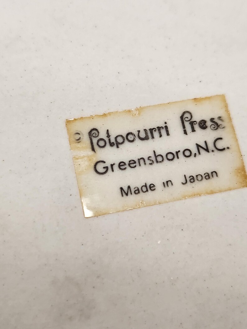 May include: Close-up of a white surface with a label that reads "Potpourri Press Greensboro, N.C. Made in Japan". The label is rectangular with a slightly aged appearance, and the text is in black.
