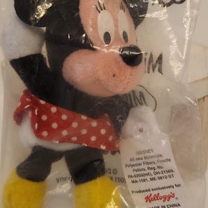 May include: A Minnie Mouse plush toy in a clear plastic bag. The toy has a red and white polka dot skirt, a red bow, and yellow shoes. The tag reads "Produced exclusively for Kellogg's".