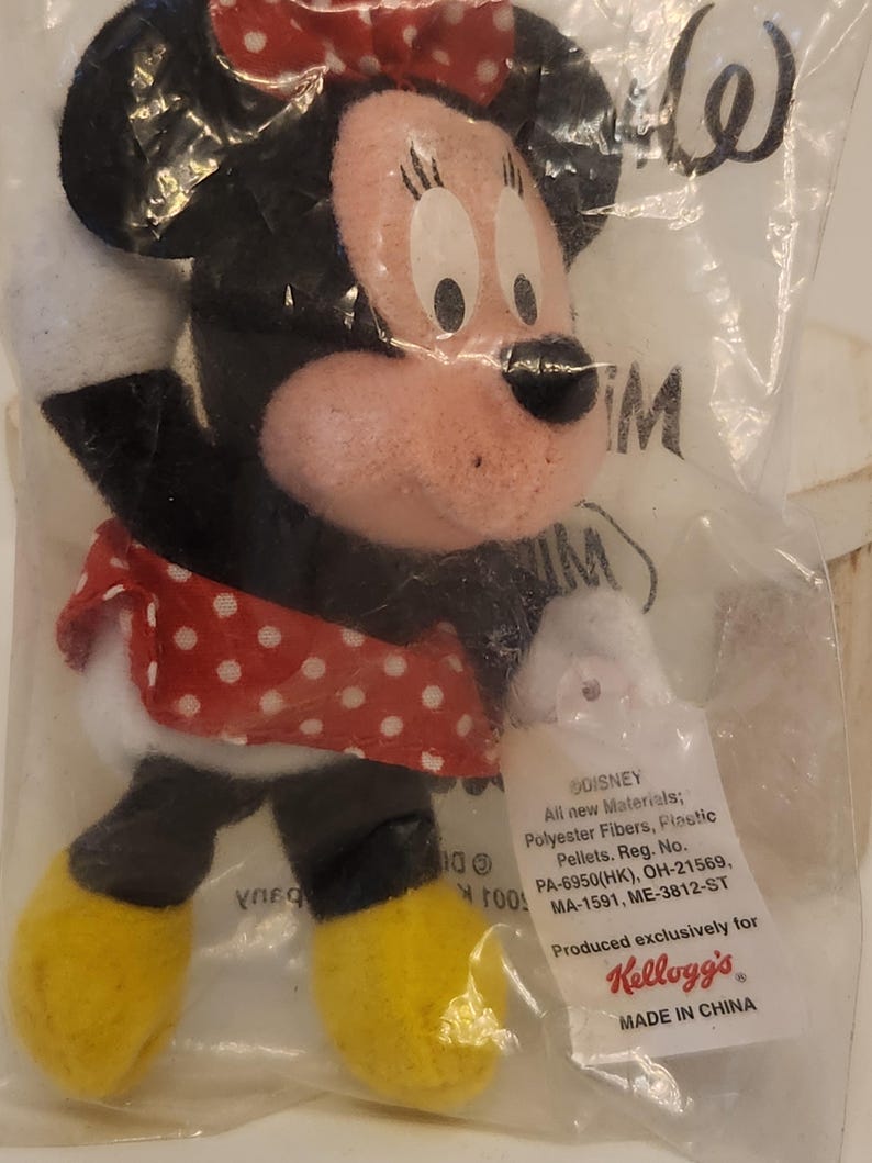 May include: A Minnie Mouse plush toy in a clear plastic bag. The toy has a red and white polka dot skirt, a red bow, and yellow shoes. The tag reads "Produced exclusively for Kellogg's".