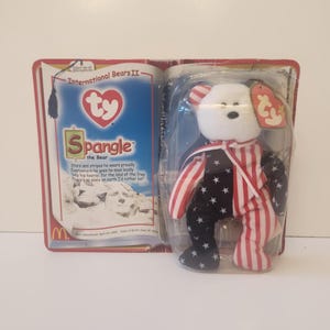 May include: A white beanie baby bear wearing a red, white, and blue outfit with stars and stripes. The bear is packaged in a plastic case with a Ty tag and a book-style cardboard backing. The book says "International Bears II" and "Spangle the Bear".