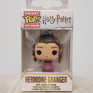 May include: A Pocket POP! Harry Potter Hermione Granger keychain figure in a clear plastic display case. The figure has brown hair and is wearing a pink dress. The packaging includes the text "HERMIONE GRANGER" and a choking hazard warning.