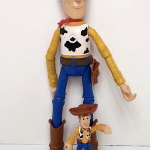 May include: Two Woody action figures, one large and one small, stand side-by-side. The larger figure is approximately 12 inches tall, wearing a yellow shirt, blue pants, and a cow-print vest. The smaller figure is about 4 inches tall, with similar attire.