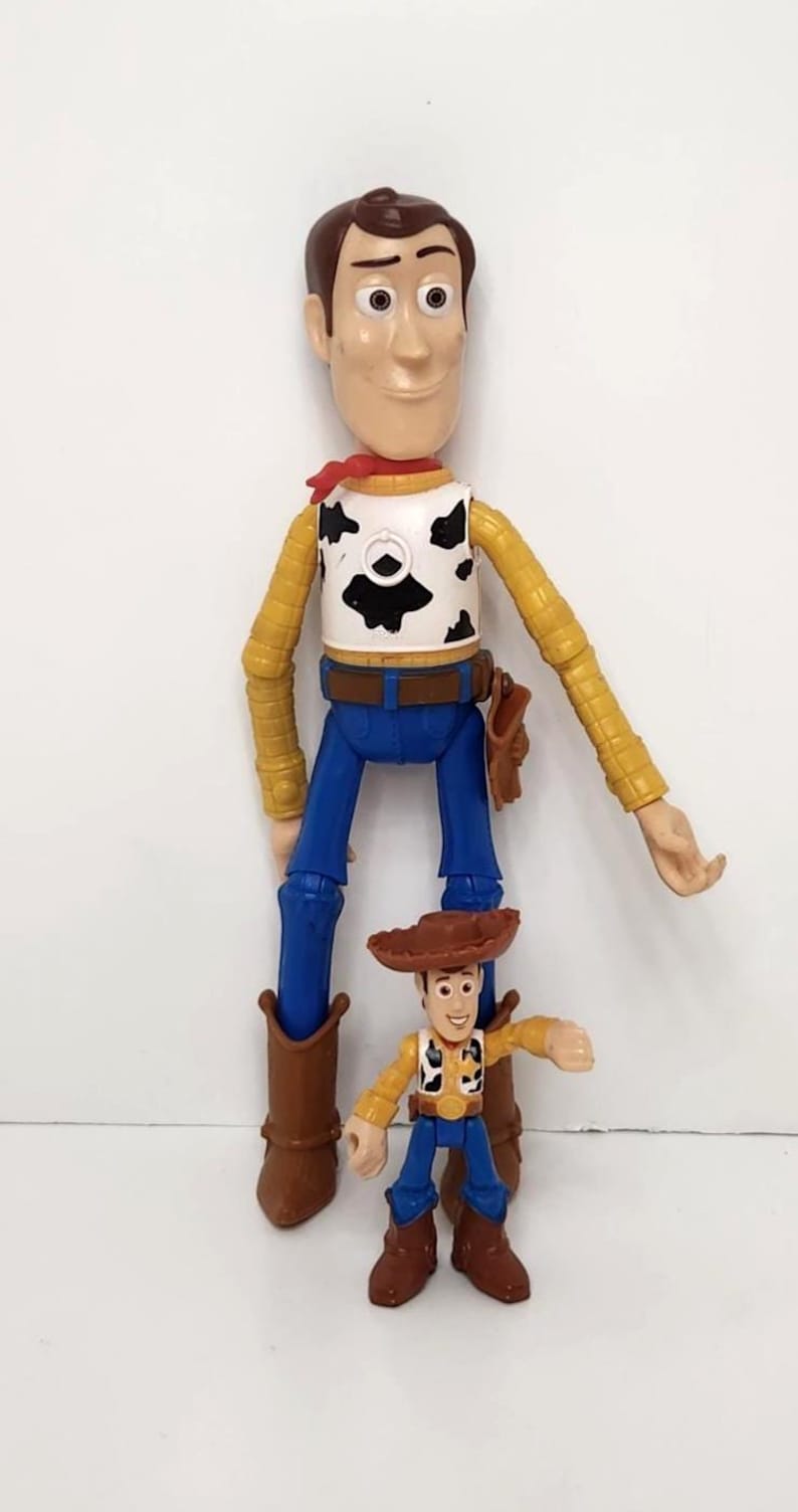 May include: Two Woody action figures, one large and one small, stand side-by-side. The larger figure is approximately 12 inches tall, wearing a yellow shirt, blue pants, and a cow-print vest. The smaller figure is about 4 inches tall, with similar attire.