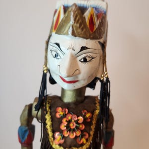 May include: A hand-carved wooden puppet with a painted face, wearing a headdress and a brown outfit with orange and yellow embellishments. The puppet has black hair and gold accents.