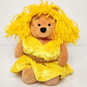 May include: A plush Winnie the Pooh toy wearing a yellow dress and a yellow yarn wig. The dress is cinched at the waist with a gold-colored rope belt. The word "Virgo" is embroidered on one foot.