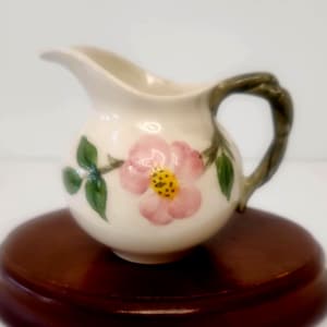 May include: A small cream pitcher made of off-white ceramic featuring a hand-painted pink flower with a yellow center and green leaves. The handle is shaped like a textured brown branch, and the pitcher has a curved spout for pouring liquids.