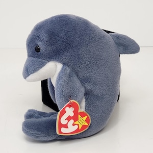 May include: A plush dolphin toy made of soft, dark gray fabric with a white underbelly and snout. The dolphin has black plastic eyes and a heart-shaped red tag attached to its side that features the white text Ty and a yellow star with Beanie Baby.