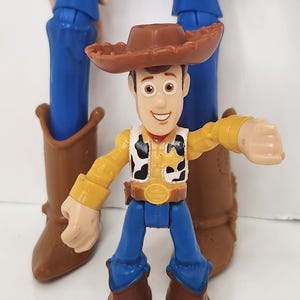 May include: A Woody action figure from the Toy Story franchise. The figure is wearing a brown cowboy hat, yellow shirt, black and white vest, blue pants, and brown boots. Another larger Woody figure is in the background.