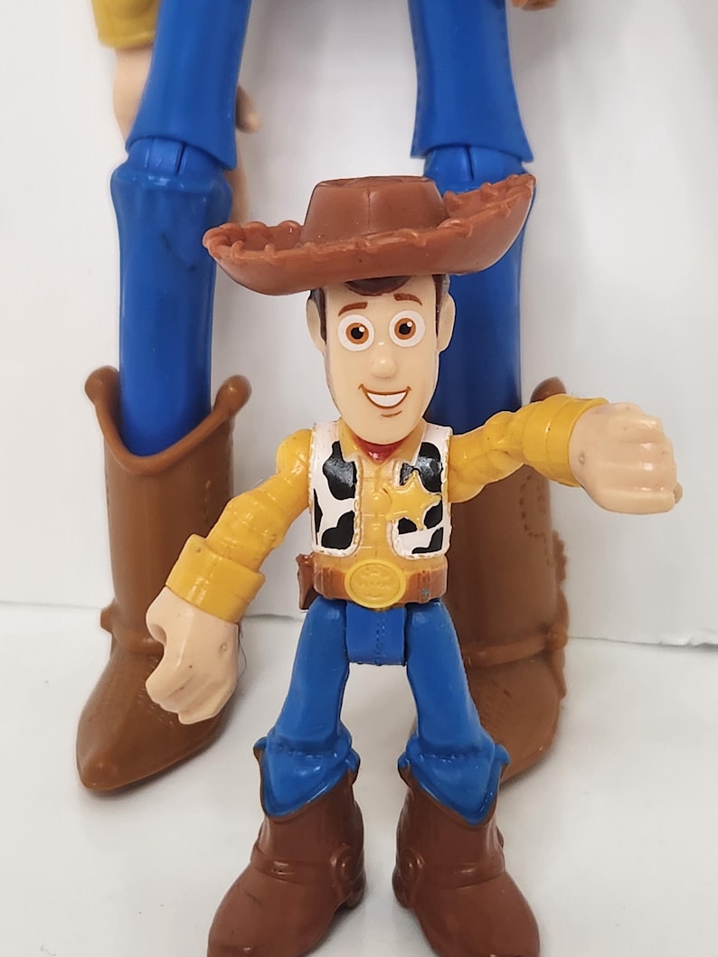 May include: A Woody action figure from the Toy Story franchise. The figure is wearing a brown cowboy hat, yellow shirt, black and white vest, blue pants, and brown boots. Another larger Woody figure is in the background.