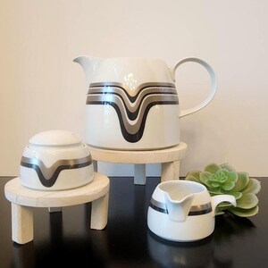 May include: A white ceramic tea set with a retro design. The set includes a large pitcher, a sugar bowl, and a creamer. Each piece features a black, silver, and brown wave pattern. The set is displayed on small wooden stands.