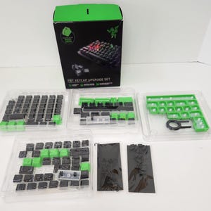 May include: A PBT keycap upgrade set with black and green keycaps, packaged in clear plastic trays. The set includes a box with the words "PBT KEYCAP UPGRADE SET" and a keyboard image. A keycap puller is also included.