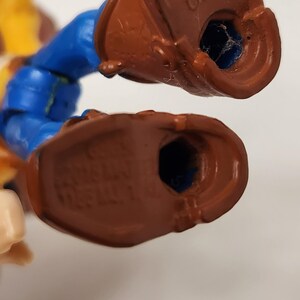 May include: Close-up of the feet of a plastic toy figure. The figure has brown boots, blue pants, and a yellow shirt. The words "MADE IN CHINA" are visible on the bottom of one boot.