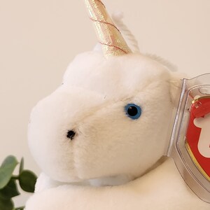 K&ouml;nnte beinhalten: Wei&szlig;es Pl&uuml;sch-Einhorn mit einem gold- und rosafarbenen, schillernden Horn und blauen Augen. Das Einhorn liegt mit dem Kopf zur Seite gedreht. Das Einhorn hat ein Ty-Etikett an seiner Seite.