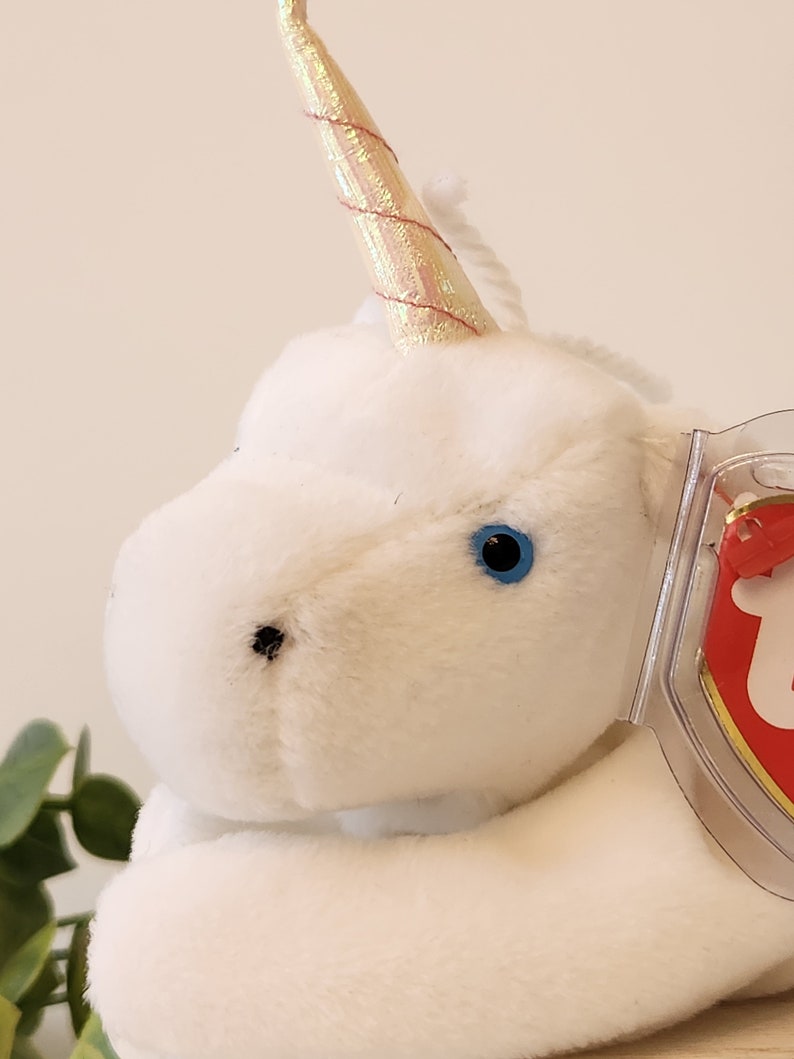 K&ouml;nnte beinhalten: Wei&szlig;es Pl&uuml;sch-Einhorn mit einem gold- und rosafarbenen, schillernden Horn und blauen Augen. Das Einhorn liegt mit dem Kopf zur Seite gedreht. Das Einhorn hat ein Ty-Etikett an seiner Seite.