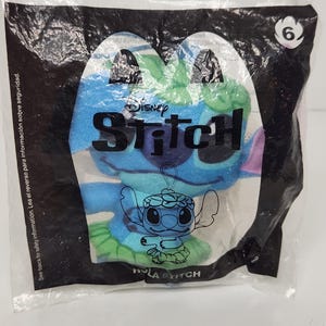 May include: A blue plush Stitch toy in a clear plastic bag. The bag has a black border and the Disney Stitch logo. The text "HOLA STITCH" is visible. A number 6 is in the upper right corner.