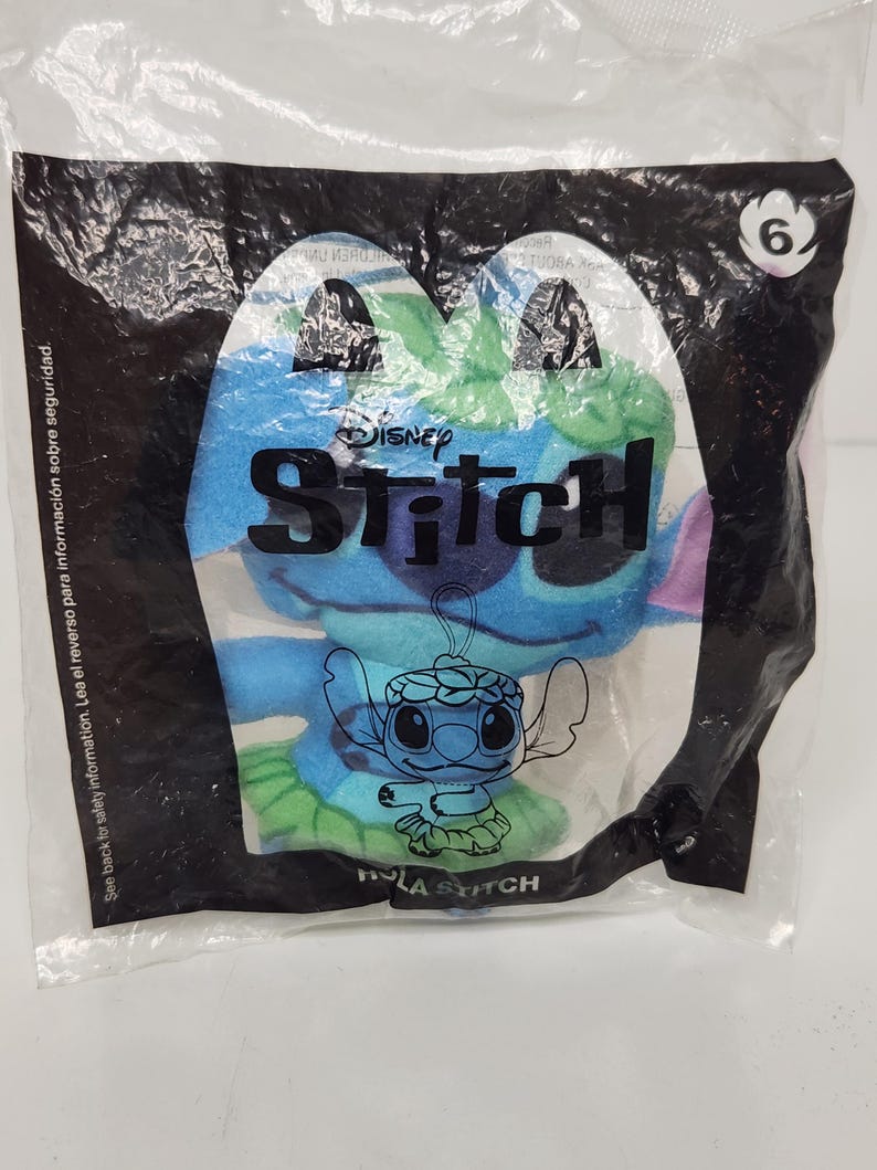 May include: A blue plush Stitch toy in a clear plastic bag. The bag has a black border and the Disney Stitch logo. The text "HOLA STITCH" is visible. A number 6 is in the upper right corner.