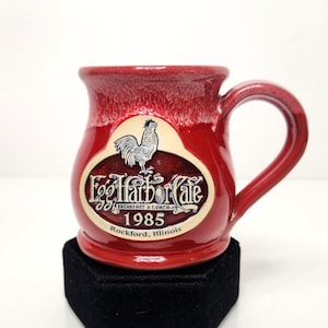 May include: A red ceramic mug with a glossy finish and a lighter red drip glaze at the rim. The front features an oval cream-colored emblem with a rooster illustration, the text Egg Harbor Cafe, Breakfast & Lunch, 1985, and Rockford, Illinois.