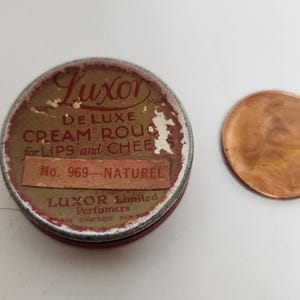 May include: Vintage Luxor Deluxe Cream Rouge tin with the text "Luxor" and "Deluxe Cream Rouge for Lips and Cheeks". The tin is round, with a red and gold color scheme. A penny is next to the tin.