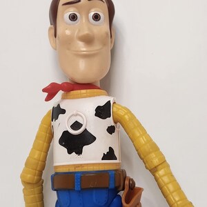 May include: A Woody action figure from the Toy Story franchise. The figure has a brown head, a red neckerchief, a white vest with black cow spots, a yellow shirt, blue pants, and a brown belt with a holster.