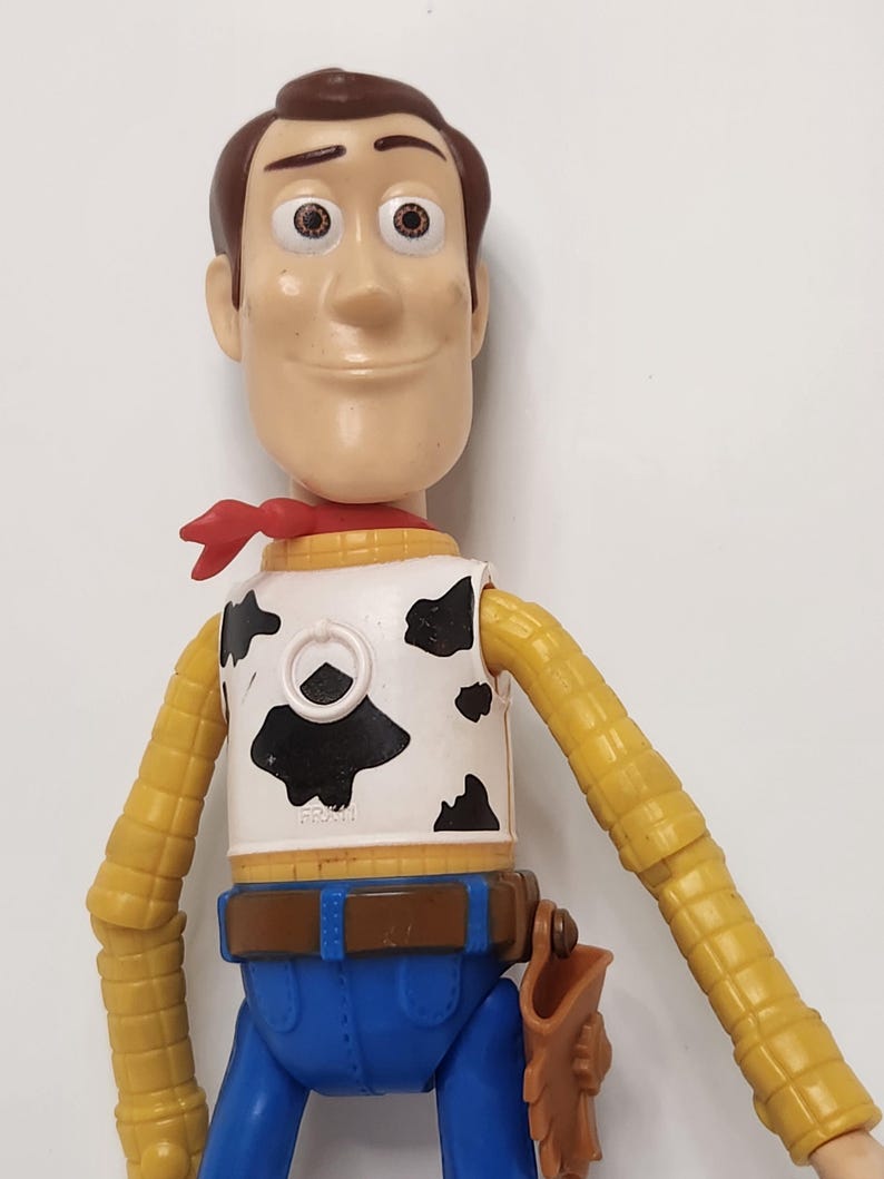 May include: A Woody action figure from the Toy Story franchise. The figure has a brown head, a red neckerchief, a white vest with black cow spots, a yellow shirt, blue pants, and a brown belt with a holster.