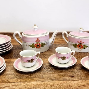 May include: A porcelain tea set featuring a teapot, a lidded sugar bowl, three cups, and six saucers. Each piece is white with a soft pink band and a hand-painted floral motif in orange and green. The set includes gold-tone trim accents.