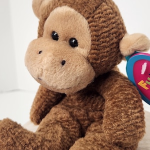 May include: A plush brown monkey with a light brown face, ears, and hands. The stuffed animal has black eyes and a tag with the word "Fun" visible. The monkey is sitting with its arms and legs extended.
